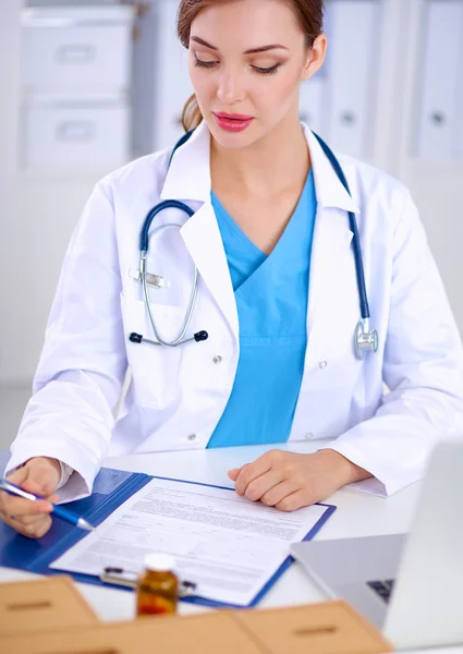 Beautiful young smiling female doctor sitting at the desk and writing.