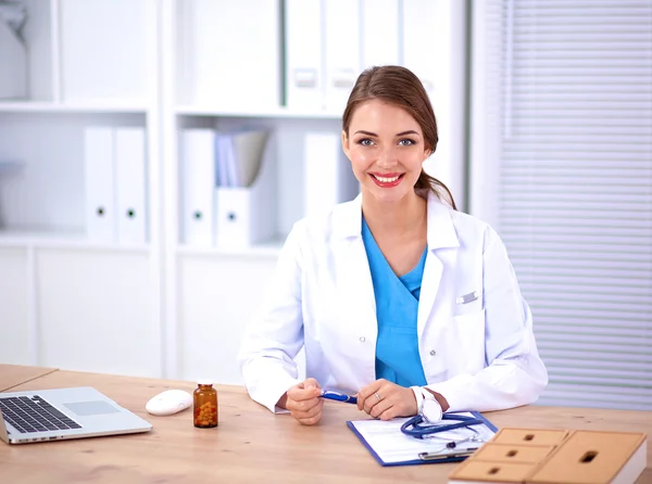 Beautiful young smiling female doctor sitting at the desk and writing.