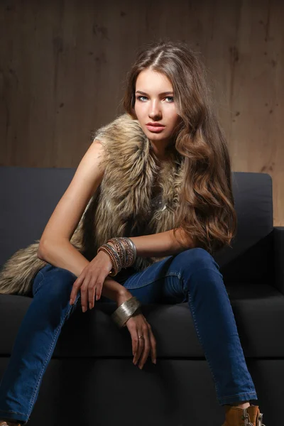 Portrait of elegant woman sitting on black sofa wearing a blue jeans and fur vest