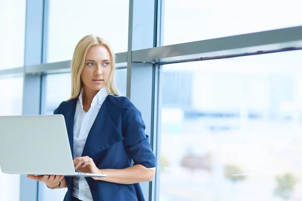 Businesswoman standing against office window holding laptop