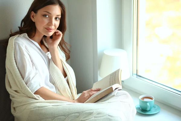 Young woman at home sitting near window relaxing in her living room reading book and drinking coffee or tea