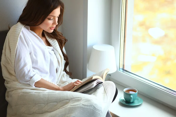 Young woman at home sitting near window relaxing in her living room reading book and drinking coffee or tea