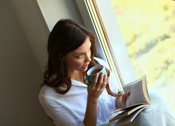 Young woman at home sitting near window relaxing in her living room reading book and drinking coffee or tea