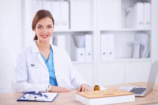 Beautiful young smiling female doctor sitting at the desk and writing.