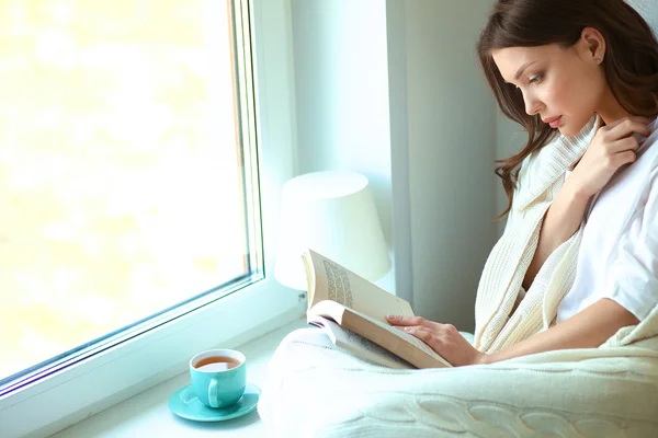 Young woman at home sitting near window relaxing in her living room reading book and drinking coffee or tea