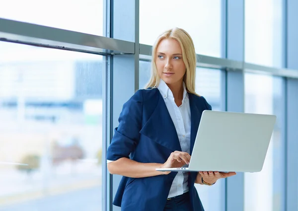 Businesswoman standing against office window holding laptop