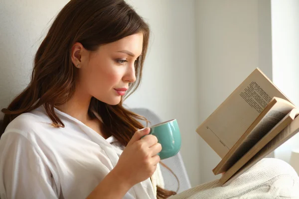 Young woman at home sitting near window relaxing in her living room reading book and drinking coffee or tea