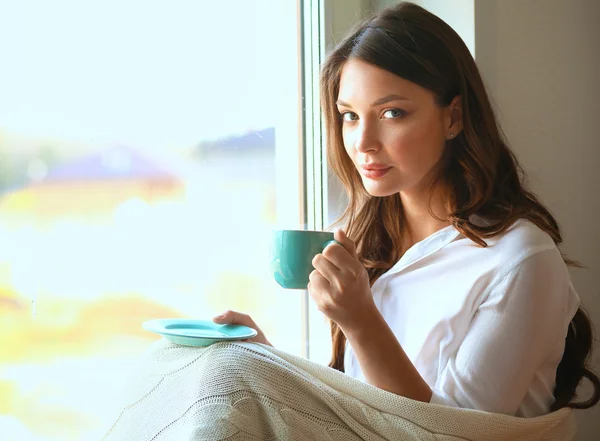 Young woman at home sitting near window relaxing in her living room reading book and drinking coffee or tea