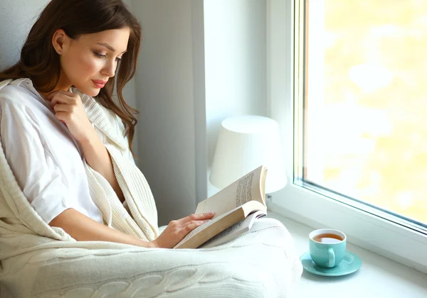 Young woman at home sitting near window relaxing in her living room reading book and drinking coffee or tea