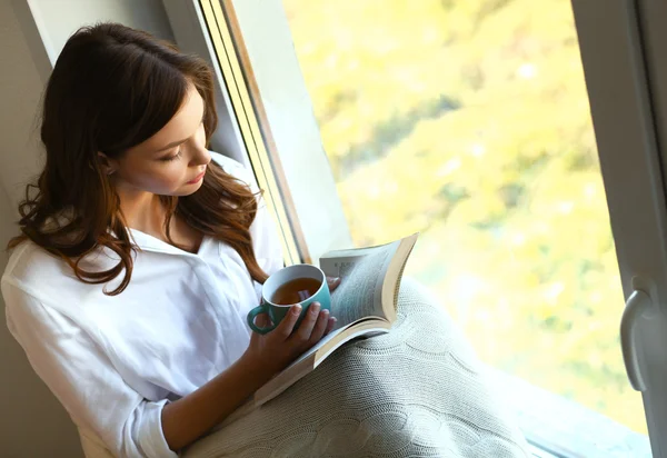 Young woman at home sitting near window relaxing in her living room reading book and drinking coffee or tea