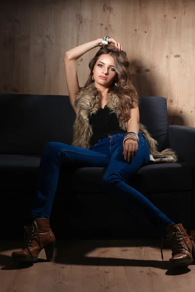 Portrait of elegant woman sitting on black sofa wearing a blue jeans and fur vest