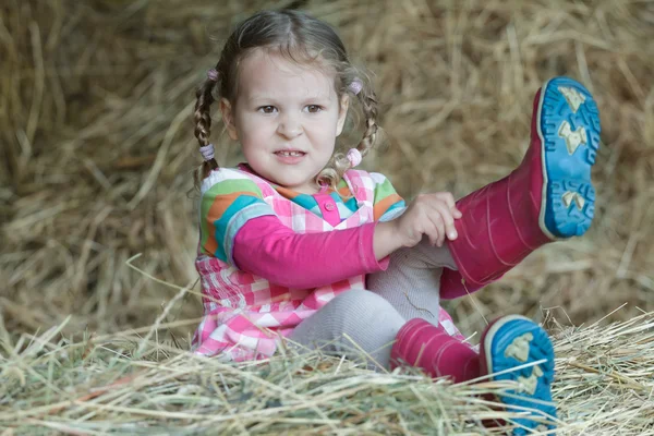 Little braided girl putting on red gum boots on dried loose grass hay in farm haystack