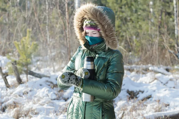 Concentrated girl wearing green shiny down coat opening stainless steel thermos flask in winter frosty wood outdoors