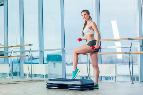 Happy young woman is doing exercises with dumbbells on step board.