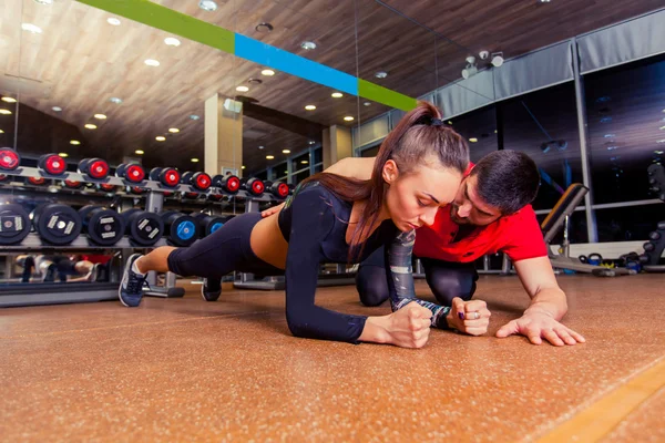 Trainer supervising a muscular woman doing plank exercises, exercise strap.
