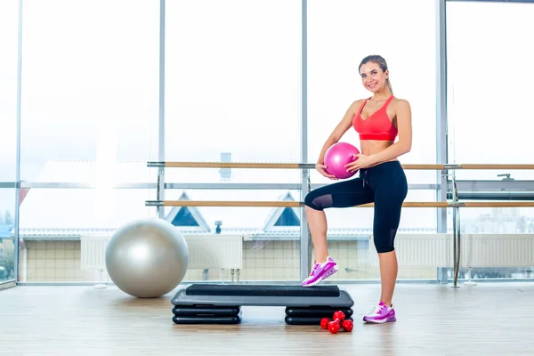 Fitness girl, wearing in sneakers, red top and black  breeches, posing on step board with ball, the sport equipment background,  gym