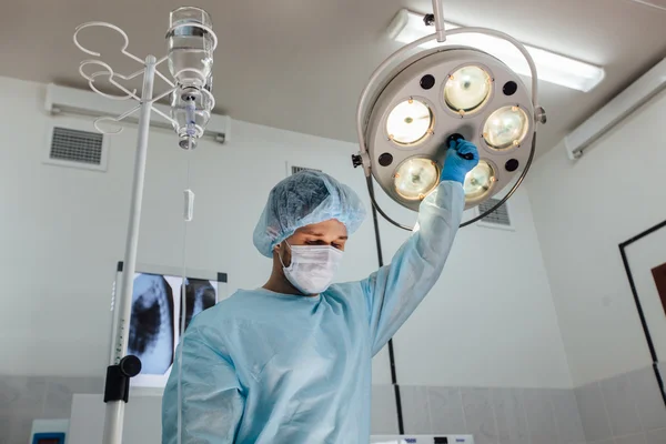 Surgery, medicine and people concept - surgeon in mask adjusting lamp  operating room at hospital