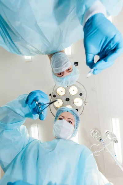 Two surgeons in protective uniform preparing for operation, looking at camera on background of surgical lamp.