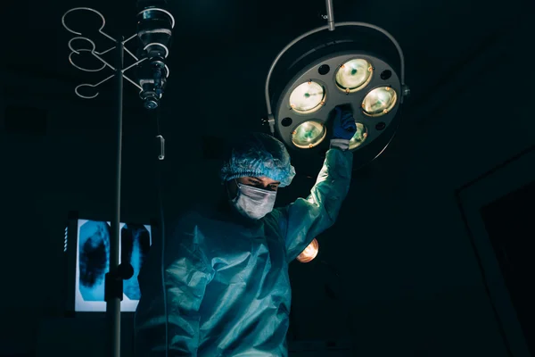 Surgery, medicine and people concept - surgeon in mask adjusting lamp  operating room at hospital