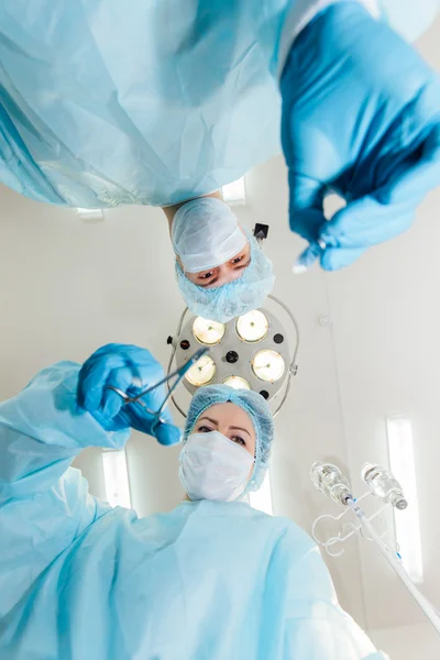 Two surgeons in protective uniform preparing for operation, looking at camera on background of surgical lamp.