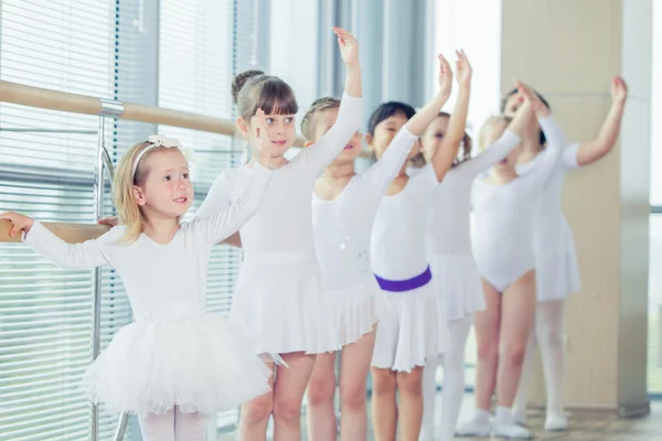 Group of seven little ballerinas standing in row and practicing ballet using stick on the wall