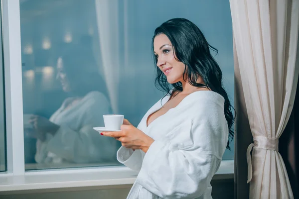 Getting warm with fresh coffee. Beautiful young woman in white bathrobe drinking coffee and looking through a window