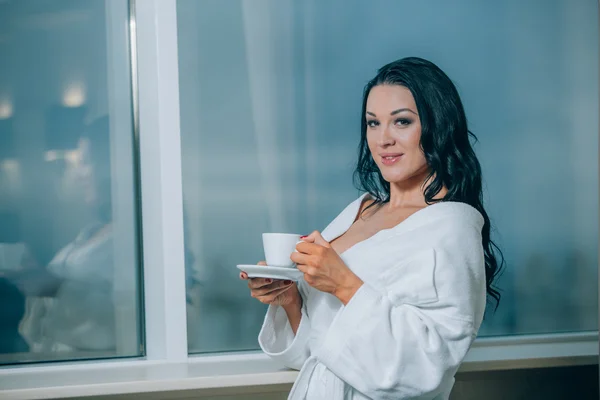 Getting warm with fresh coffee. Beautiful young woman in white bathrobe drinking coffee and looking through a window