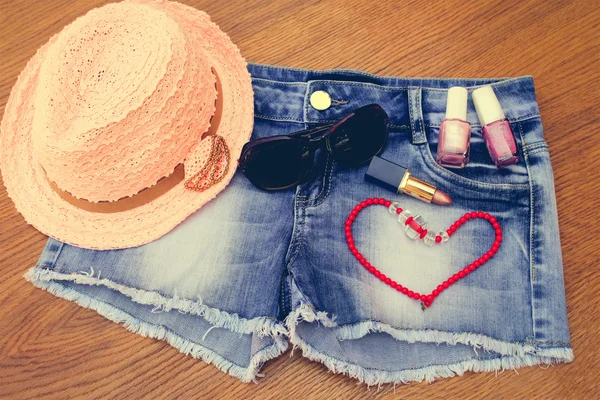 Summer women's accessories: red sunglasses , beads, denim shorts, sun hat, nail polish, lipstick open. Toned image.