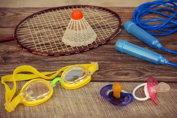 Pacifier and sports equipment: the birdie is on the racket, skipping rope, swimming goggles on wooden background. Concept of sports to be engaged with early childhood.