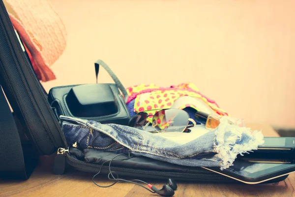 Open the suitcase with tourist things: women's hat, swimsuit, camera, denim shorts, dresses, sunglasses, perfumes, nail polish, mobile phone, tablet on wooden background.