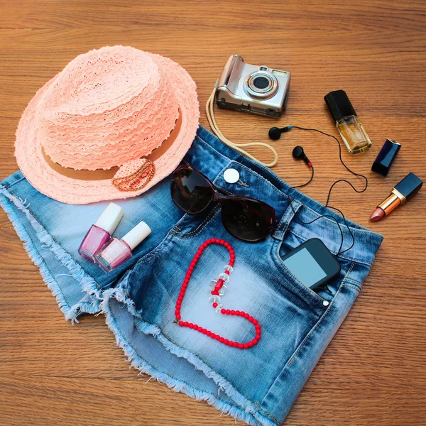 Summer women's accessories: red sunglasses, beads, denim shorts, mobile phone, headphones, a sun hat, camera, nail polish, open lipstick, perfum,. Toned image