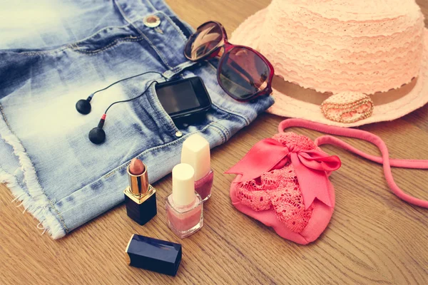 Summer women's accessories: sunglasses, beads, denim shorts, mobile phone, headphones, a sun hat, handbag, lipstick, nail polish. Toned image