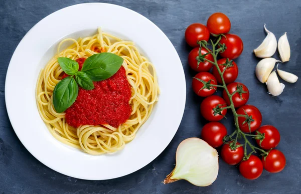 Spaghetti with marinara sauce and basil leaves on top, decorated with cherry tomatoes, garlic, onion on blue background.