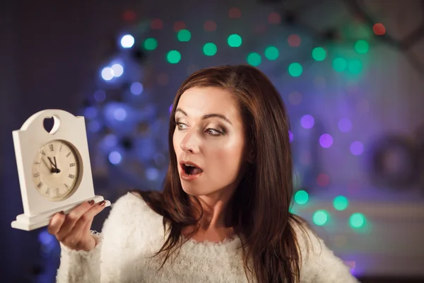 Attractive surprised woman holding clock near christmas tree