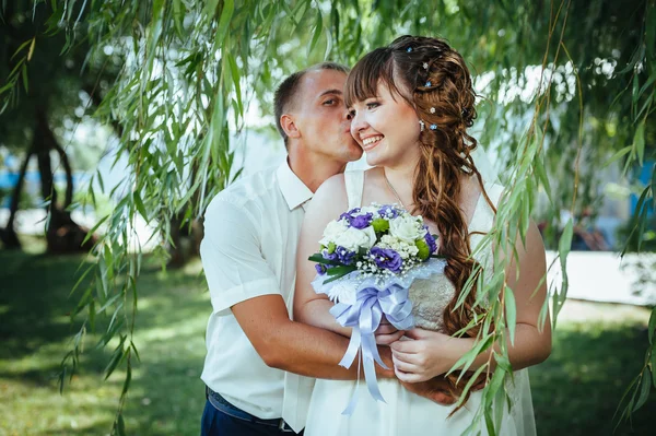 Wedding couple kissing in green summer park. bride and groom kissing, standing together outdoors, hugging among green trees. Bride holding wedding bouquet of flowers