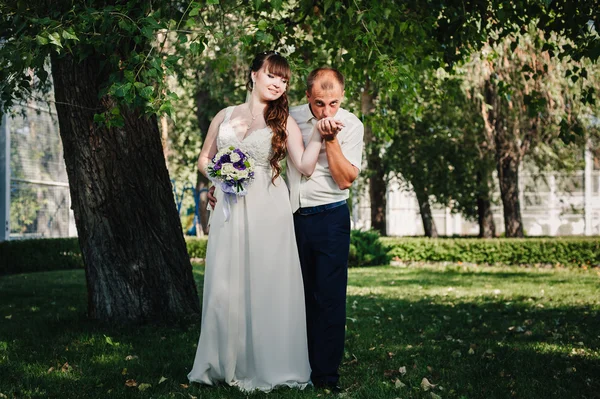 Wedding couple kissing in green summer park. bride and groom kissing, standing together outdoors, hugging among green trees. Bride holding wedding bouquet of flowers