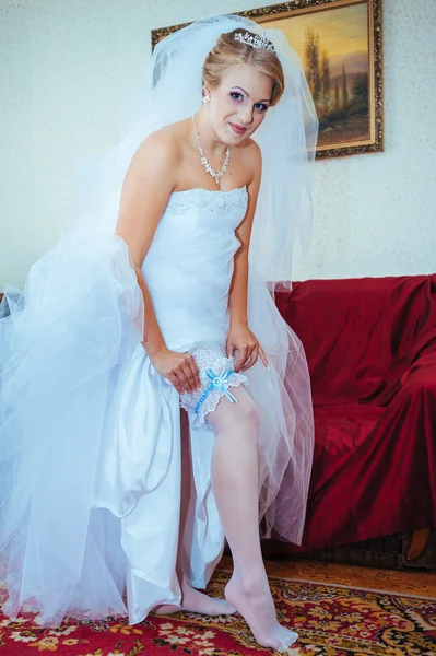 Garter on leg of bride, slim sexy bride in bridal luxury dress showing her silk garter with golden ribbon. woman have final preparation for wedding ceremony. Wedding day moments