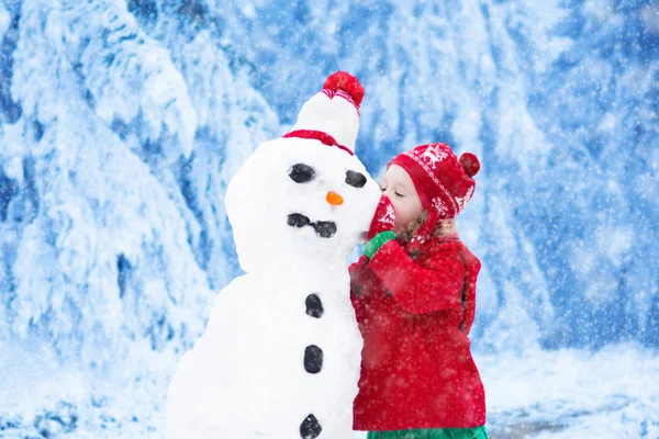 Funny little toddler girl in a red knitted Nordic hat and warm coat playing with a snow man. Kids play outdoors in winter. Children having fun at Christmas time. Child building snowman at Xmas.