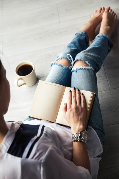 Young modern girl in torn jeans reading a book with a big cup of