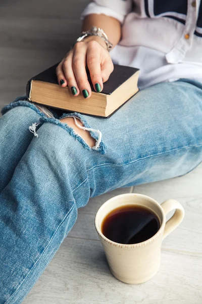 Young modern girl in torn jeans reading a book with a big cup of coffee. Fashion, lifestyle, lifestyle, recreation, education, hobbies.