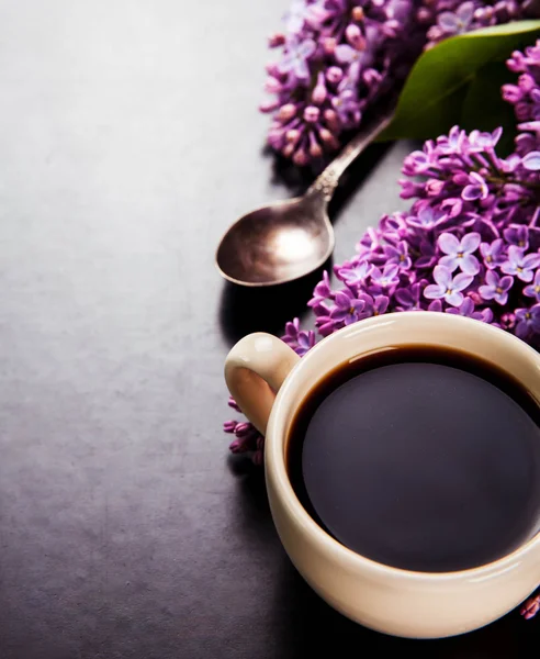 Black coffee in a cup, a spoon and fresh lilac flowers on black background