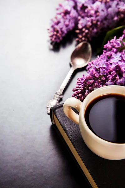 Black coffee in a cup, book, a spoon and fresh lilac flowers on black background