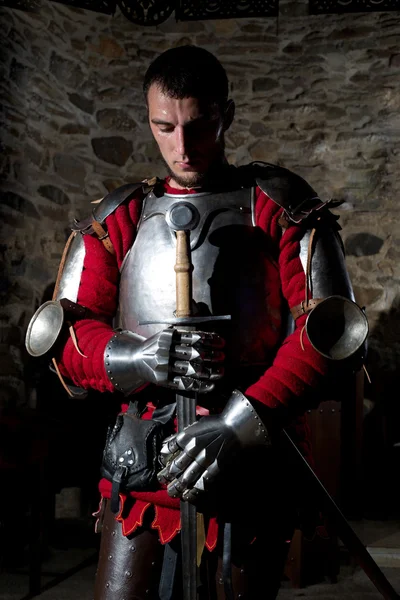 Knight Standing With Head Bowed in Prayer and Holding Metal Sword Against Old Stone Wall