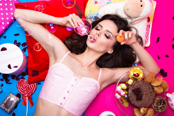 Beautiful fresh girl doll with soap bubbles lying on bright backgrounds surrounded by sweets, cosmetics and gifts. Fashion beauty style.