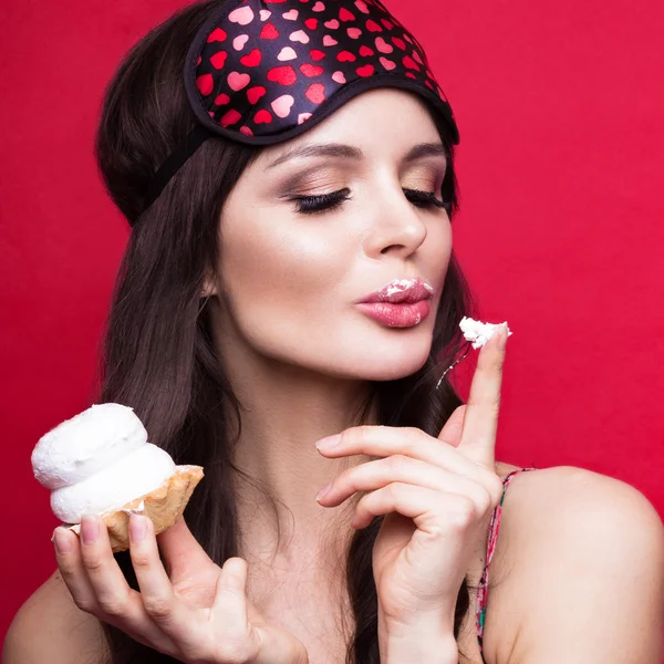 Funny young woman in sleeping mask and pajamas, sweets on red background. beauty face.