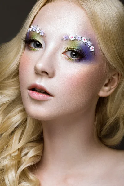 Beautiful young blonde woman with creative make-up color, curls and flowers on eyebrows. Beauty face. Art makeup.