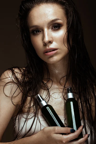 Beautiful girl with a bright make-up, wet hair and skin, bottle of cosmetic products in hands. Beauty face.