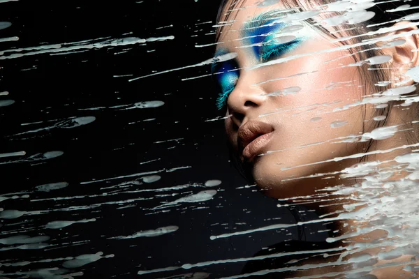 Beautiful Asian girl with bright blue make-up behind glass and drops of wax. Beauty face.