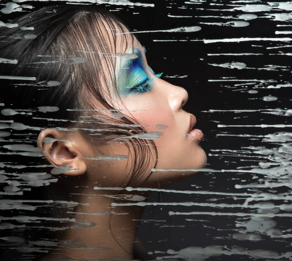 Beautiful Asian girl with bright blue make-up behind glass and drops of wax. Beauty face.