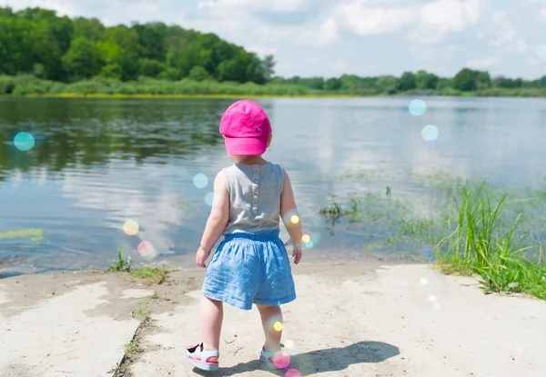 Little girl in pink cap costs on bank of lake.